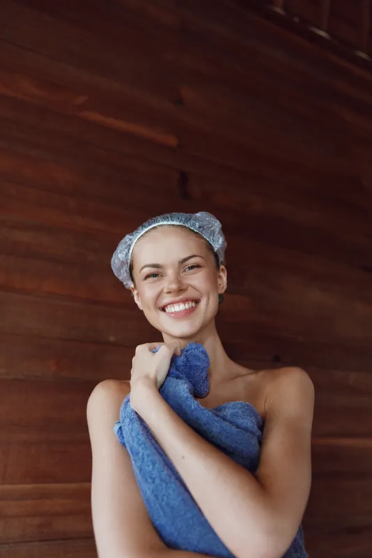 Smiling woman wrapped in a blue towel enjoying spa relaxation with a hair cap indoors against wooden walls, radiating happiness and comfort during self-care.