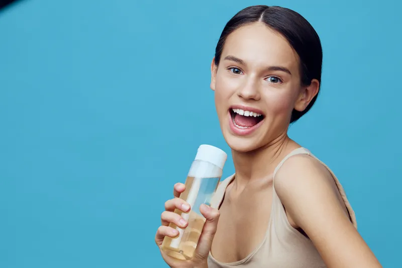 Happy young girl holding a skincare bottle, smiling brightly against a blue background, showcasing joy and healthy skin care practices
