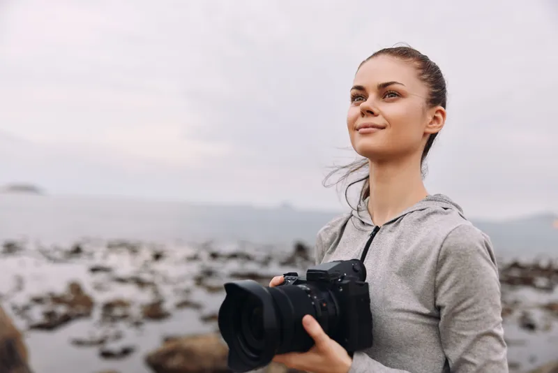 Young woman with a camera by the seaside, wearing a gray hoodie, showcasing joy and passion for photography against a serene coastal background