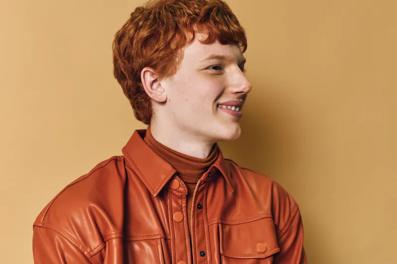 Young man with red hair smiling and looking to side wearing brown leather jacket and sweater on beige studio background. Casual fashion portrait with natural expression.