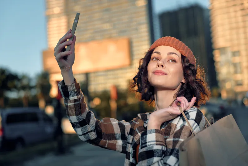 Woman selfie shopping urban golden candid captures authenticity and joyful mindful living as a young shopper poses with bags, emotional storytelling and warm golden hour glow in city streets.