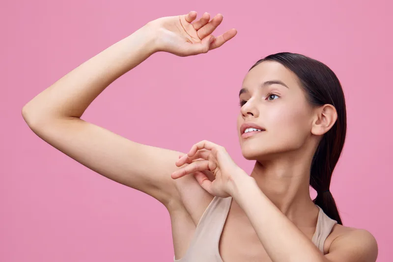 Young woman with smooth skin posing elegantly in a light tank top against a pink background, showcasing confidence and grace