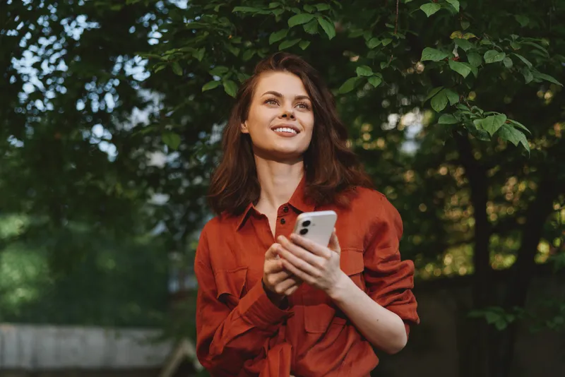 Smiling woman in a red shirt holding a smartphone outdoors. Leafy background with soft sunlight. Concept of casual lifestyle, connection, and everyday moments