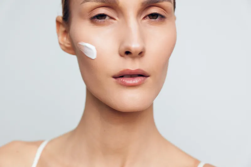 Closeup portrait of woman with clear skin applying white cream on cheek, neutral expression, minimal makeup, soft lighting, plain background, skincare routine concept