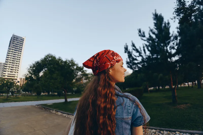 Lifestyle woman in modern boho-western denim street style with red bandana and long hair, captured in natural park setting with warm film color tones.