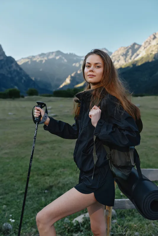 A young woman hiker stands in an alpine meadow with distant mountains behind her, posing with a trekking pole and a rolled mat, ready for adventure and exploration.