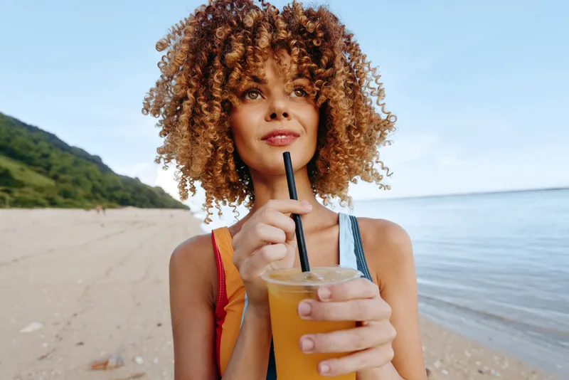 Woman with curly hair on beach, holding a drink with straw, summer pointing gesture, relaxed sunny outdoor scene, relaxation and wellness concept, ocean shoreline, natural daylight, in focus