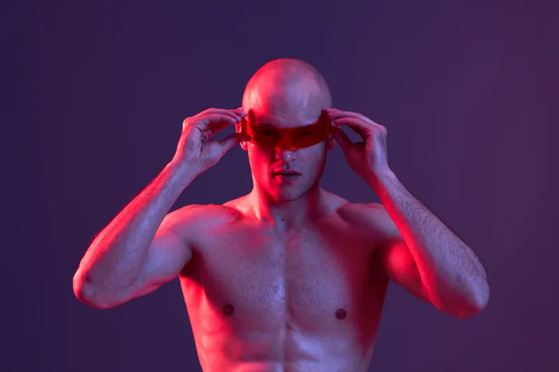 Athletic man wearing red goggles, showcasing a bold look in a vibrant atmosphere with purple lighting and dramatic contrasts, emphasizing strength and confidence.