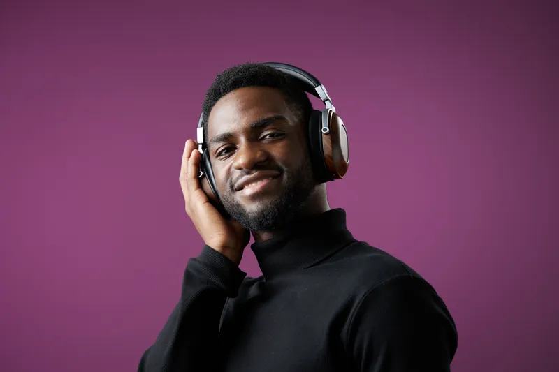 Young African man enjoying music with headphones, smiling against a purple background, showcasing a modern and relaxed style