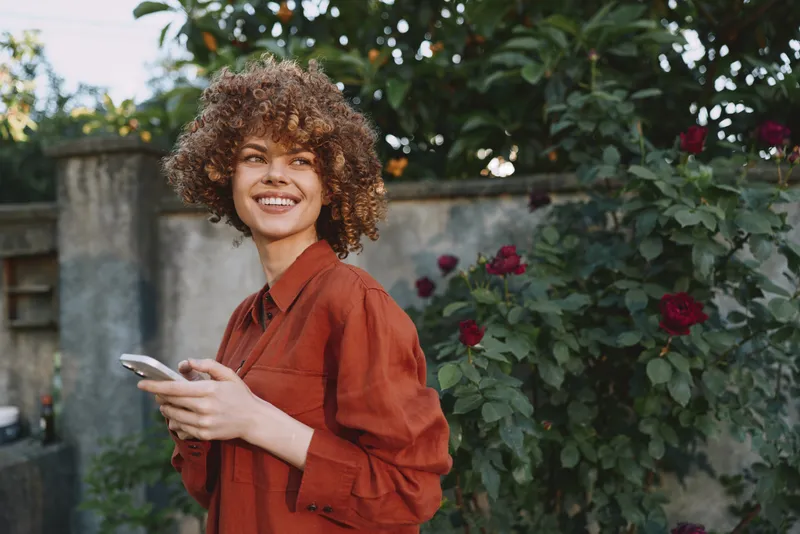 Joyful woman in an earthy shirt enjoys an outdoor moment, phone in hand, bright garden background, candid lifestyle portrait capturing spontaneous happiness, warmth, and connection