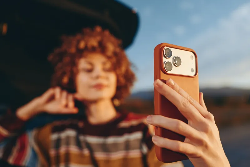 Woman lifestyle portrait outdoors, holding smartphone for a selfie, hand near face, curly hair under hat, street urban background with soft bokeh, casual sweater, bright daylight