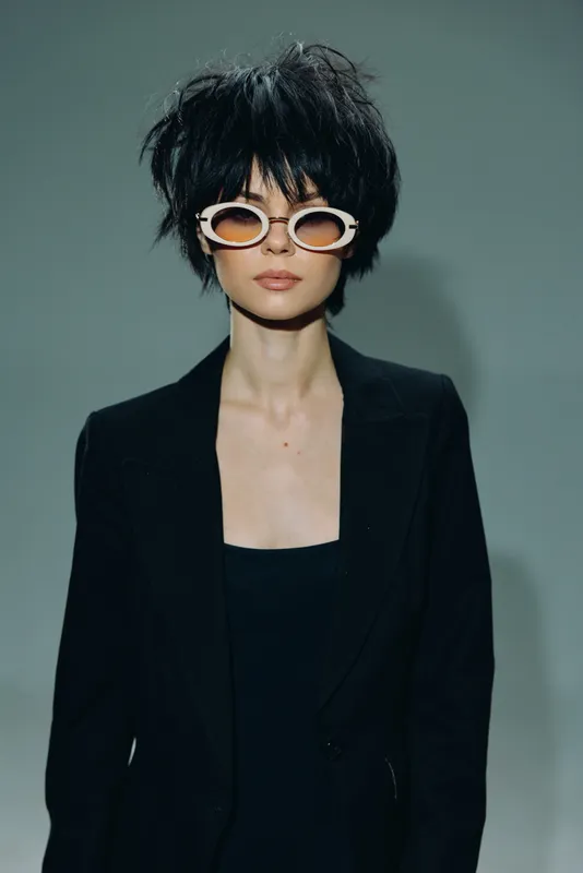 Stylish woman with short black hair wearing fashionable sunglasses and a black blazer against a gray background