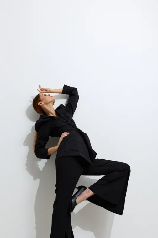 Fashion woman posing in black suit against white wall, showcasing stylish clothing, confident posture, and modern minimalist aesthetic with dramatic shadow play and sleek footwear.