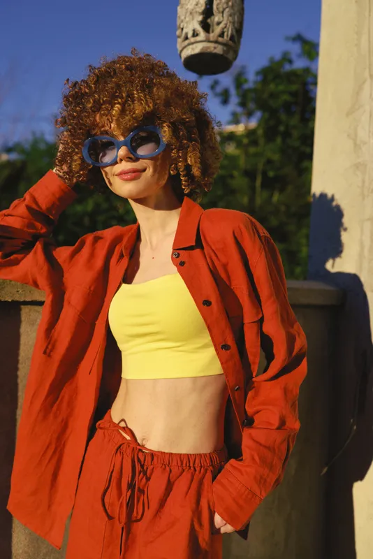 Young woman with curly hair poses outdoors in a bright red jacket and yellow crop top, blue sunglasses, relaxed stance, radiating confidence and sunny street style