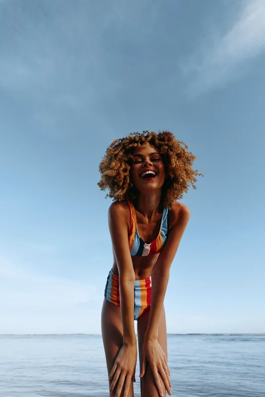 Young woman with curly hair in a striped swimsuit laughs on a sunny beach today