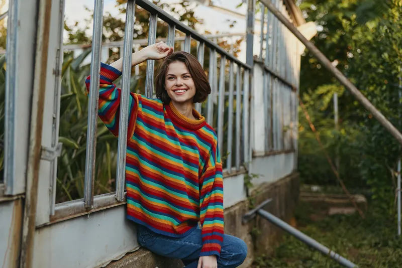 Woman in a colorful striped sweater leaning against a greenhouse window, smiling casually. Young woman in casual outfit and jeans, relaxed outdoor lifestyle portrait with cozy autumn mood.