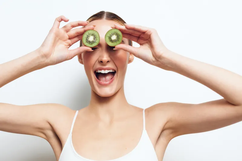 Young woman with long hair holds two kiwi slices over her eyes and smiles joyfully against a white background in a playful and fresh concept of healthy lifestyle and fun.