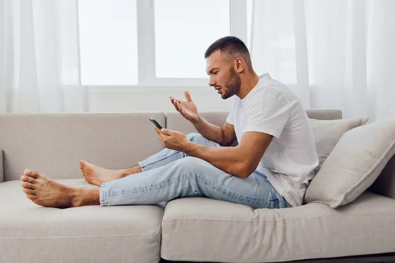 Frustrated man sitting on a couch, using smartphone, expressing emotions, relaxed home environment, light colors, soft sunlight streaming through windows.