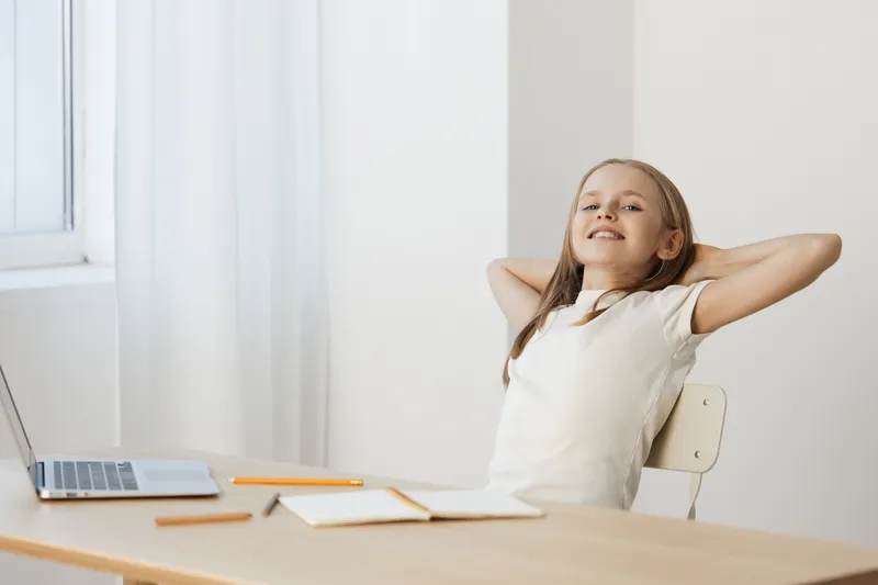 Happy girl relaxing at a desk with a laptop, showcasing a positive attitude towards work and study, in a bright and minimalist setting
