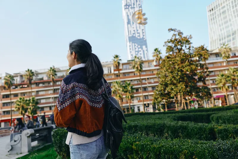 Woman with backpack walks through a sunny city park, modern buildings in the background, green hedges and palm trees, candid street scene, casual fashion in urban exploration