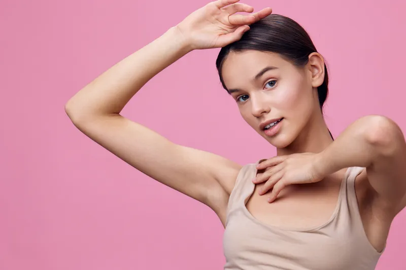 A young woman with a natural expression poses against a soft pink background, showcasing a minimalist aesthetic and neutral sleeveless top