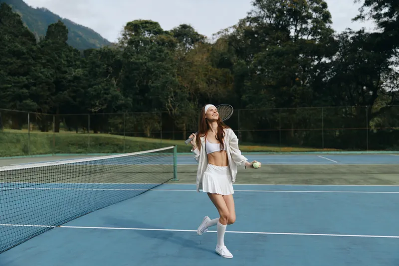 Joyful young female tennis player in white attire celebrating on a sunny outdoor court, showcasing athleticism and enthusiasm with a green nature backdrop
