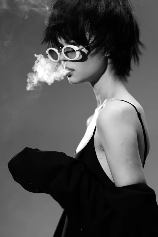 Black and white portrait of a stylish woman with short hair blowing smoke and wearing round sunglasses and a jacket