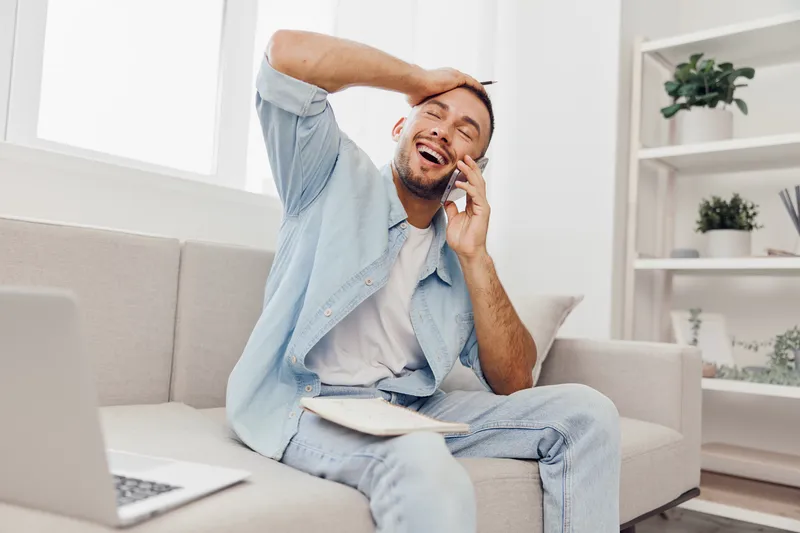 Joyful man engaged in an emotional phone call, blending work and personal life, expressing vulnerability and embracing authentic imperfection in a cozy living space.