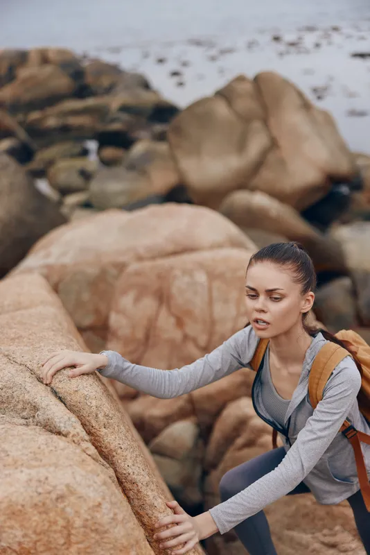 Young woman climbing rocks by the coastline, showcasing determination and outdoor adventure in a relaxed outfit with a backpack