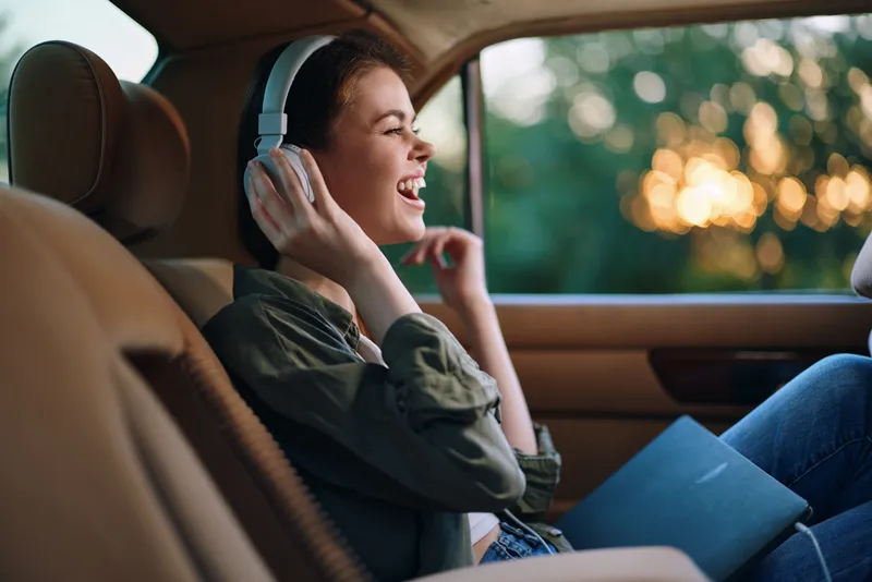 Young woman enjoying music in a car during sunset, wearing casual clothing, with headphones on and a joyful expression, surrounded by a warm color palette.