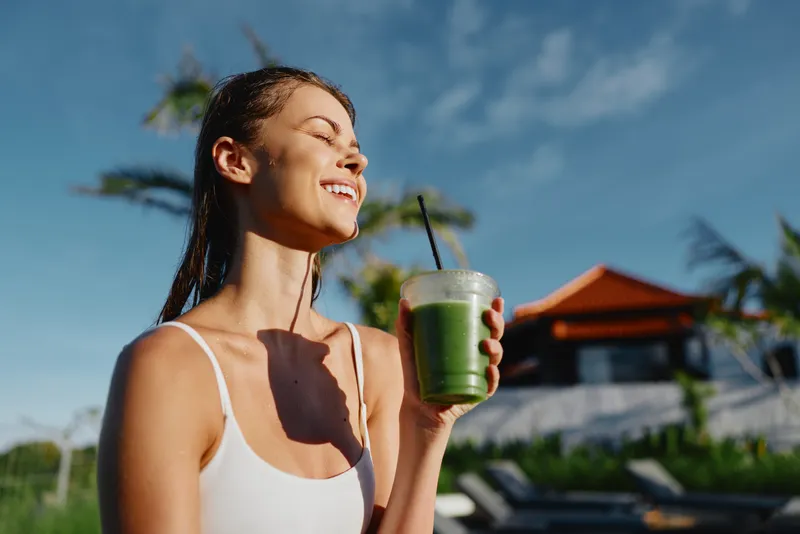 A young woman in a white tank top enjoys a green smoothie by a sunlit poolside, smiling with eyes closed amid palm trees, capturing a relaxed, healthy lifestyle moment.