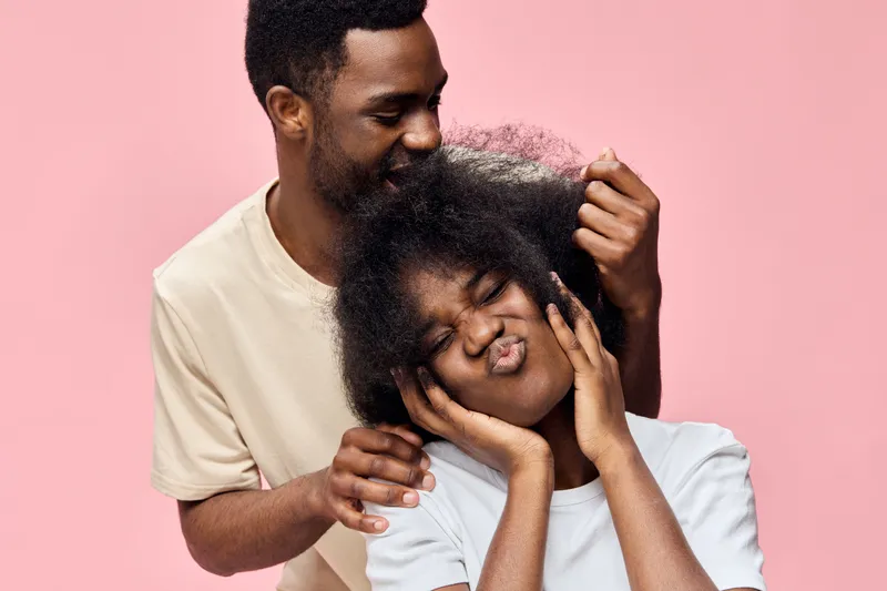 A joyful couple playfully interacting with natural hair against a pastel pink background, showcasing fun, love, and self expression in a vibrant, seasonal setting