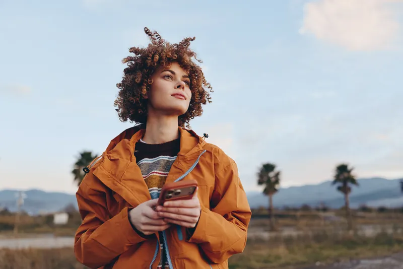 Woman with curly hair in orange jacket holding a smartphone outdoors under a blue sky, lifestyle moment for communication and travel, soft light, casual stance, nature background
