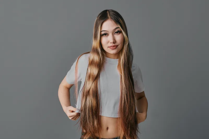 Young woman with long straight hair wearing cropped t shirt looks at camera with confident expression on gray background. Portrait of female beauty and casual style.