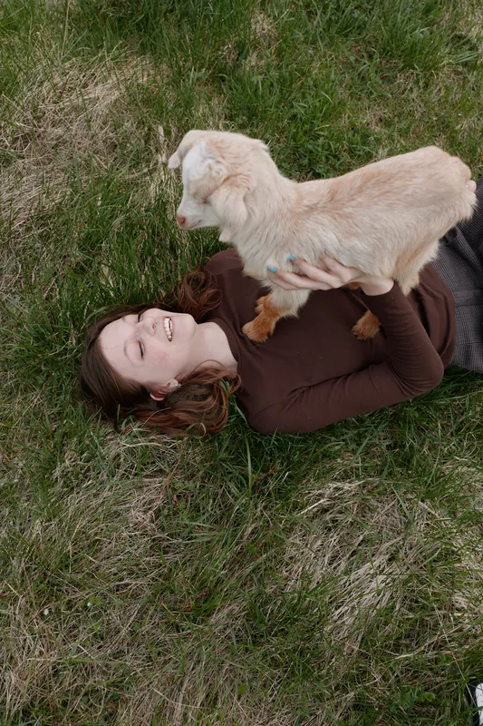 Young woman lies on green grass holding a baby goat above her with a joyful smile in a relaxed outdoor lifestyle moment during spring.