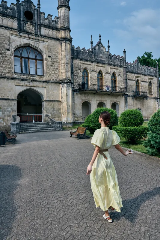 Young woman in a pale yellow dress walks across a gravel courtyard toward a grand historic castle, capturing a serene travel moment with architectural charm and soft light.