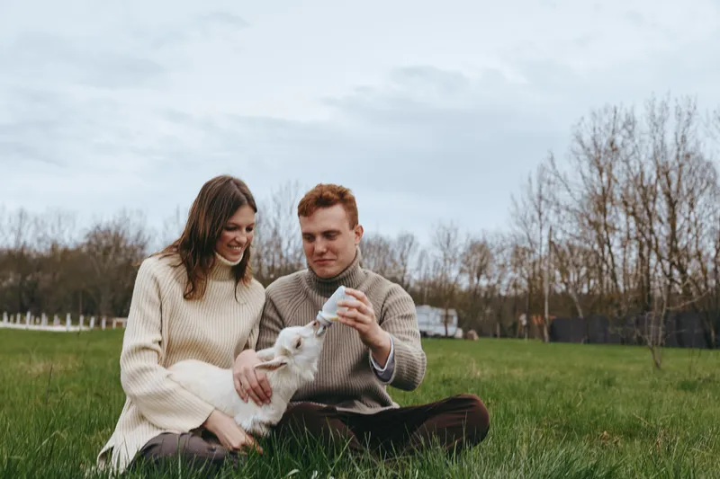 A young couple in warm sweaters sits on grass feeding a baby goat with a bottle, enjoying a peaceful lifestyle moment outdoors in early spring.