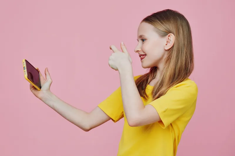 A cheerful young girl taking a selfie with a smartphone against a vibrant pink background, wearing a bright yellow t shirt, conveying happiness and positivity