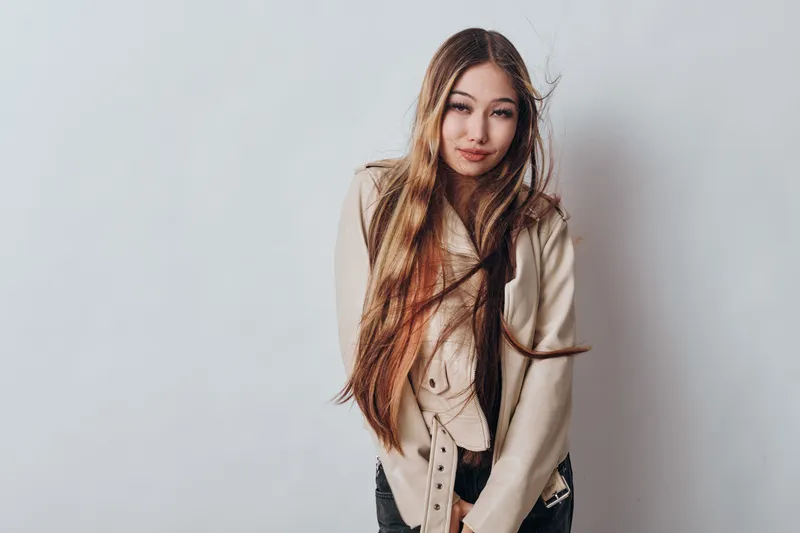 Young woman with long hair wearing beige jacket posing against white background. Casual style, natural makeup, and relaxed expression in modern portrait photography.