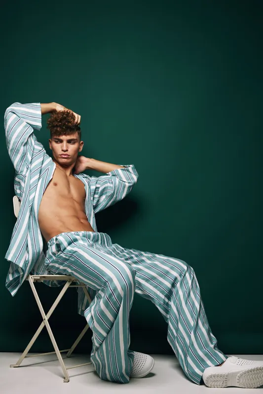 Young man with curly hair wearing striped pajamas sitting on chair with hands behind head against dark green background in studio.