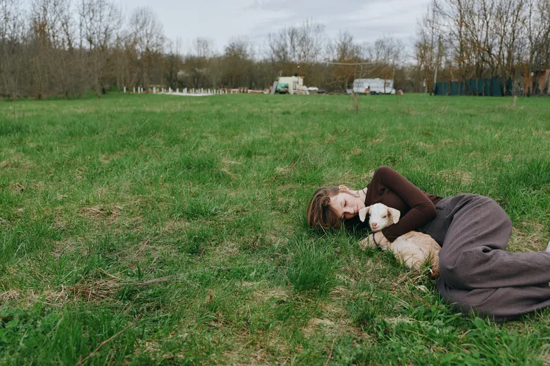 Casual outdoor scene with a woman lying on lush grass, tenderly embracing a fluffy goat kid, sunlit moment, relaxed mood, gentle breeze, countryside setting, earthy tones, peaceful companionship