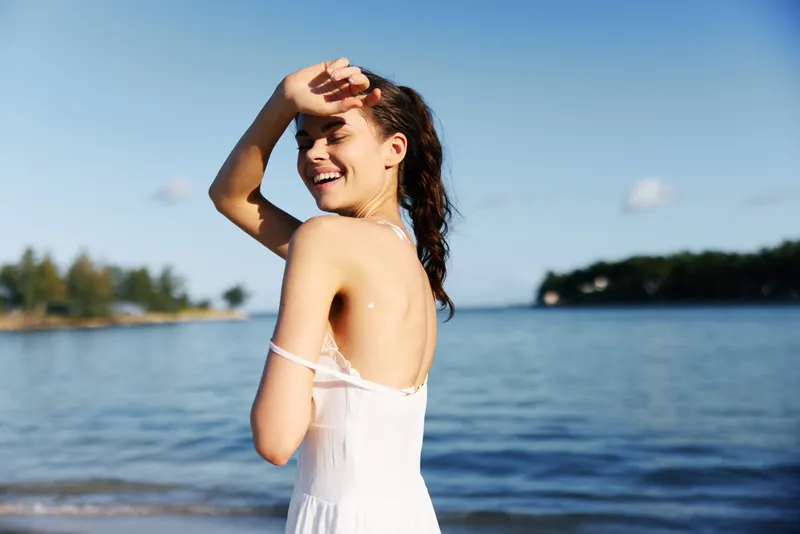 A joyful woman stands on a sunny beach, arms raised in a carefree pose, wearing a light white dress as gentle waves wash onto golden sand.