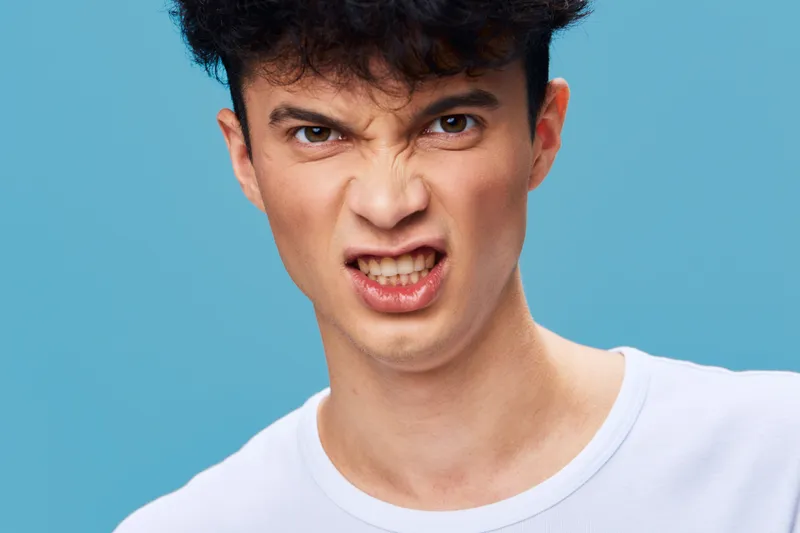 Angry man with curly hair wearing a white shirt on blue background expressing frustration and tension in a close-up portrait, young adult male emotion.