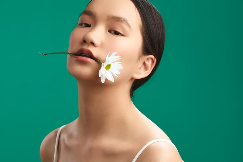 asian woman with a white flower held between lips against a vivid green backdrop, creating a fresh editorial portrait with soft lighting and calm, poetic mood.