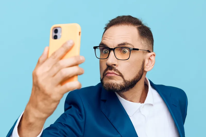 A business professional man in a blue suit takes a selfie against a solid blue background, conveying focus, confidence, and curiosity in a clean corporate portrait.