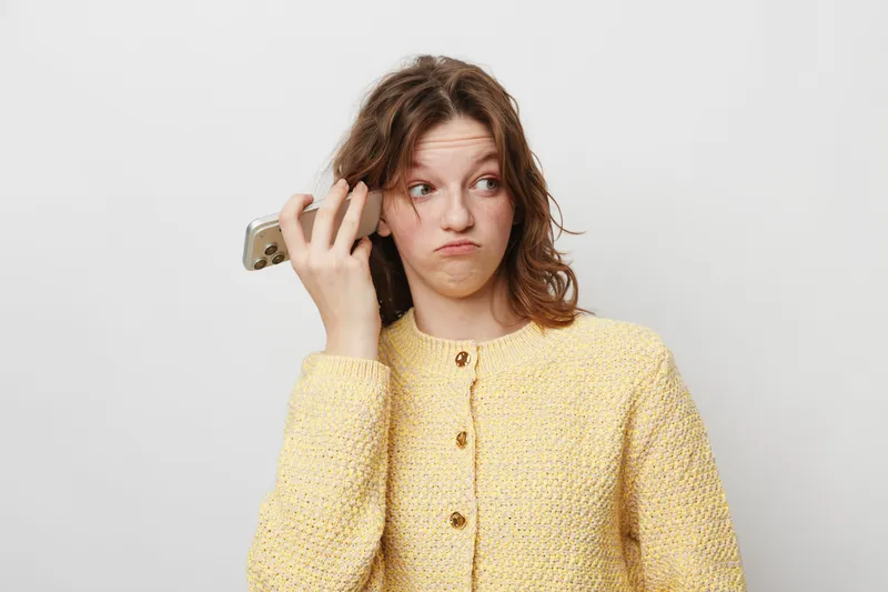 Young woman in yellow sweater holds smartphone near ear with confused expression. Isolated on white background. Concept of misunderstanding, communication, and technology frustration.