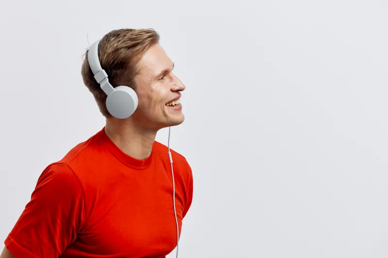 Smiling man wearing red shirt listening to music with large headphones on plain white background studio portrait happiness joy leisure lifestyle concept