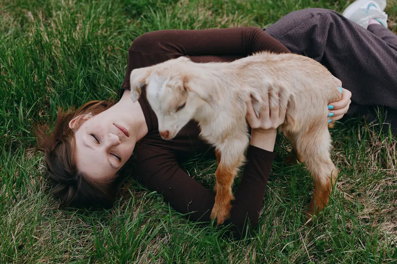 Woman lies on grass hugging a goat kid, enjoying a peaceful outdoor moment, warm light, casual lifestyle, gentle affection, bond with animal, serene mood and connection.