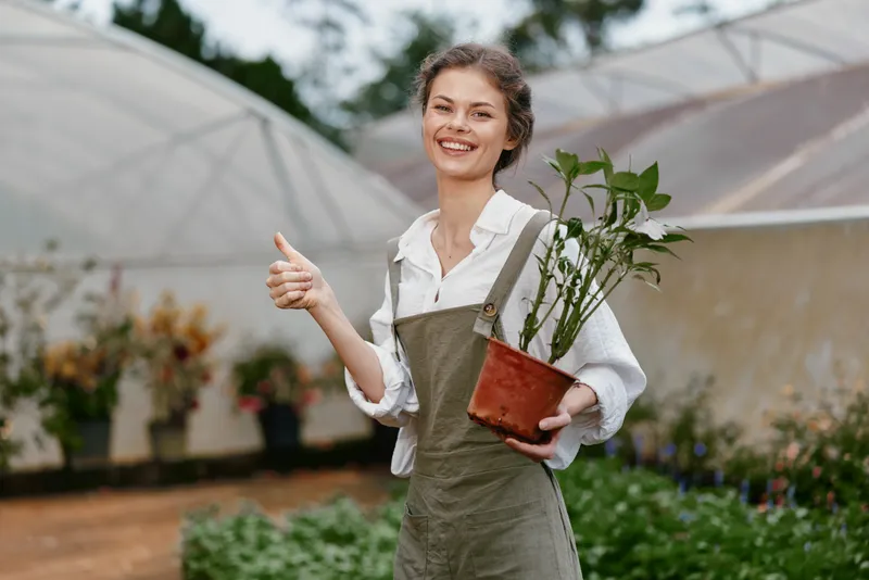 Happy woman in overalls holding a potted plant in a greenhouse, showcasing healthy gardening and a connection with nature in a vibrant setting