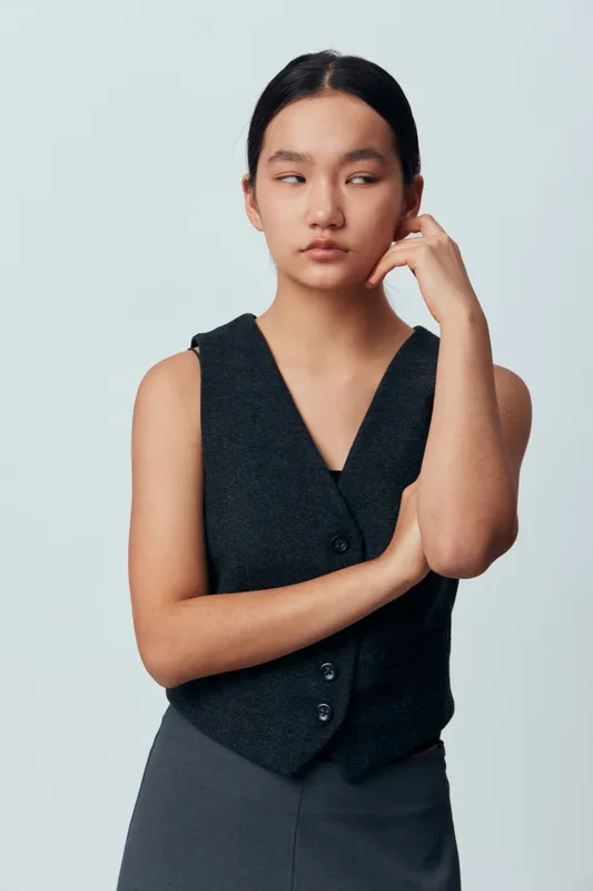 Young woman with serious expression standing with arms crossed and hand near face wearing dark sleeveless vest and skirt on plain light background in studio portrait.